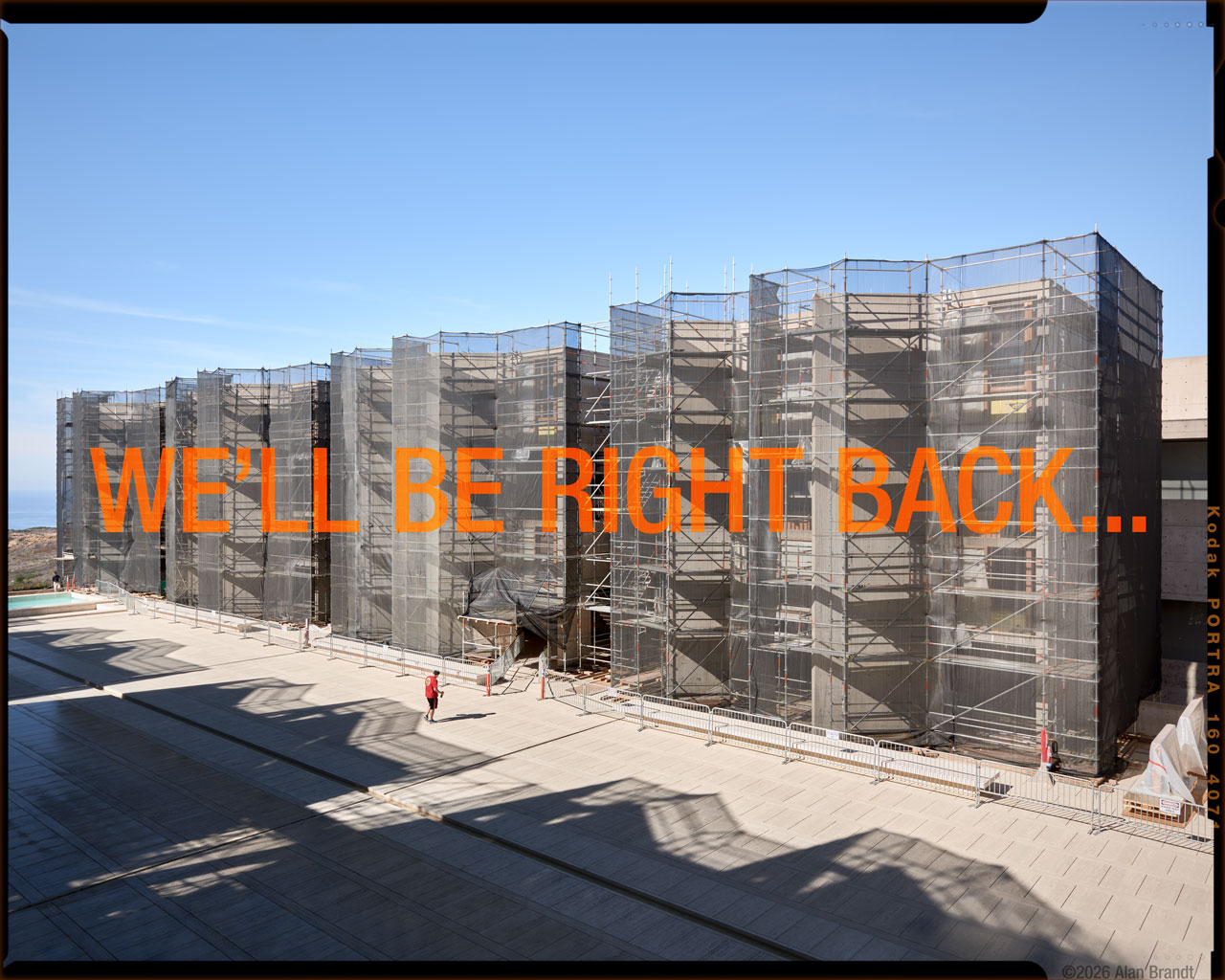 Exterior architectural photo of Salk Institute with scaffolding during renovation. Foreground has shadows from more scaffolding out of view. Shot on large format 4x5 film. Overlay text reads ”We'll be right back" to use image as a placeholder while website for Alan Brandt Architectural Photography is being updated.